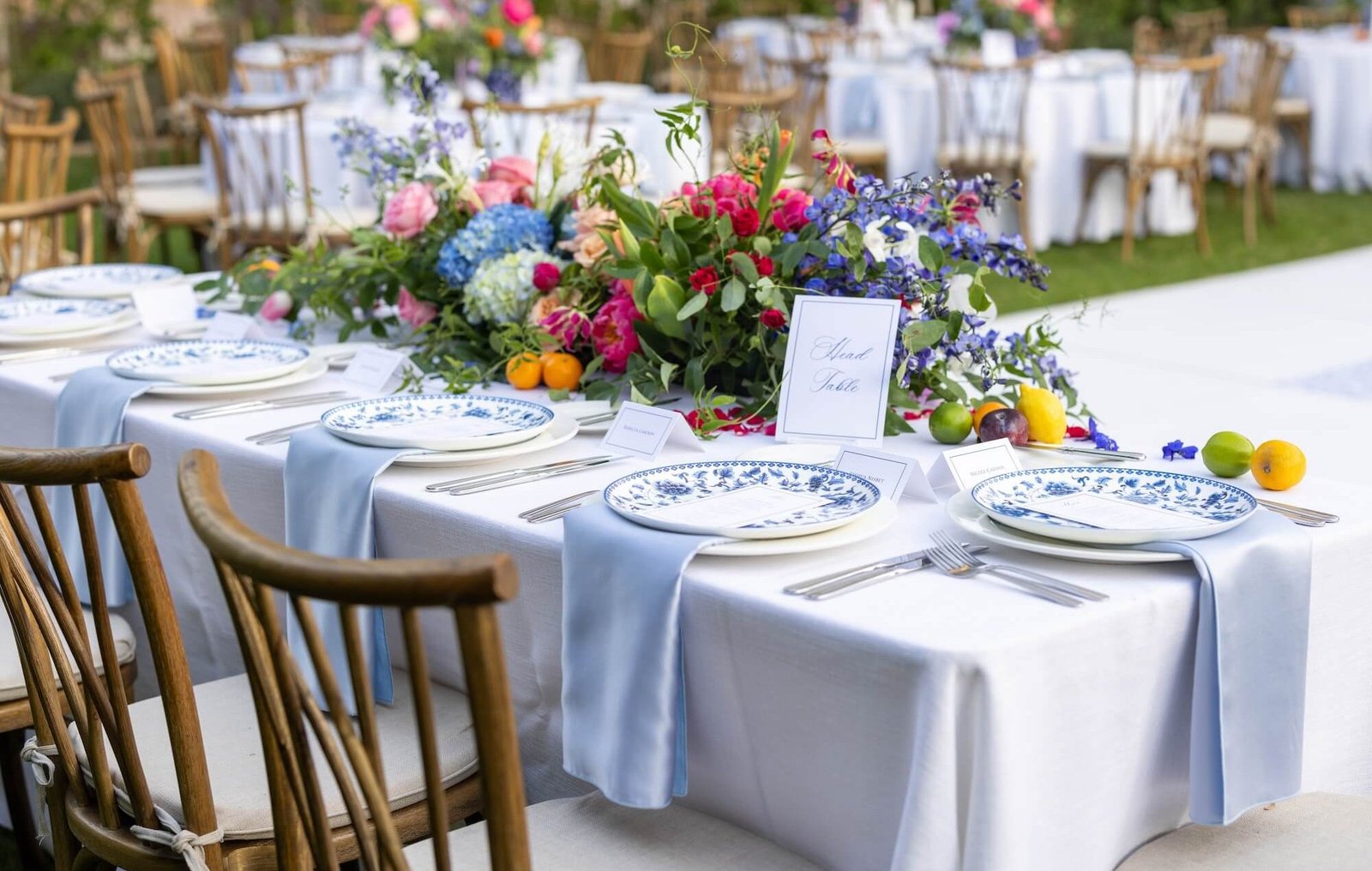 Outdoor engagement reception table set with crisp white linens, blue-and-white patterned plates, pastel floral centerpiece, fresh fruit accents, and wooden chairs — perfect for engagement catering services near me