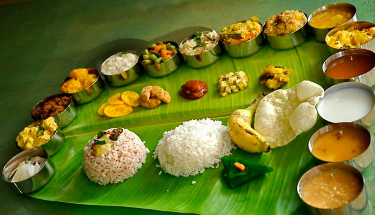 South Indian housewarming catering menu on a banana leaf featuring steamed rice, curd rice, banana, papad, and an array of small bowls with sambar, rasam, curries, vegetable sides, chutneys, and sweets